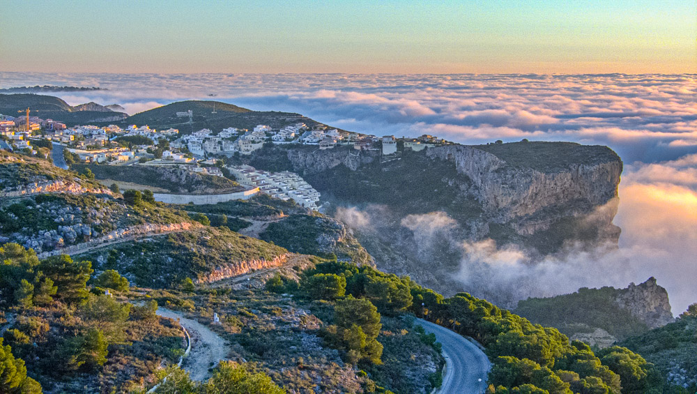 Vue panoramique des falaises de Cumbre del Sol surplombant une mer de nuages au coucher du soleil, illustrant le relief côtier et les quartiers résidentiels.