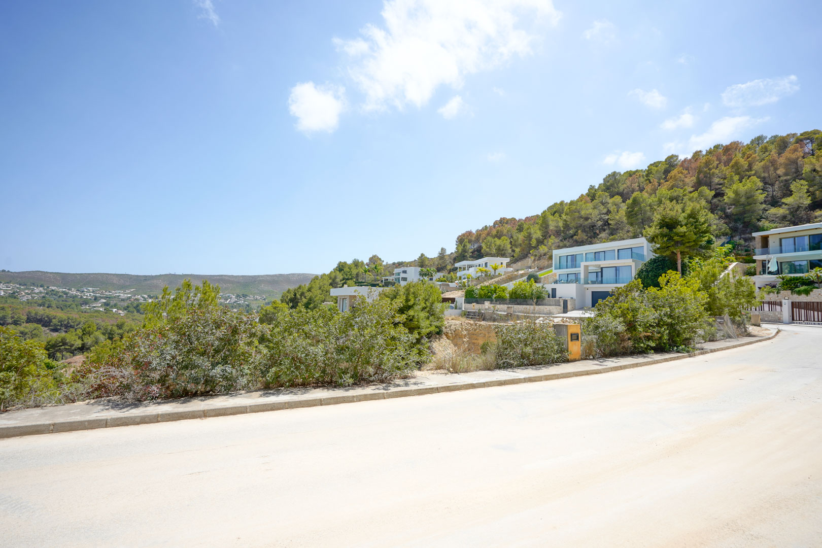 Rue pavée dans un lotissement moderne, présentant des villas contemporaines nichées à flanc de colline et entourées d'une forêt de pins méditerranéenne.