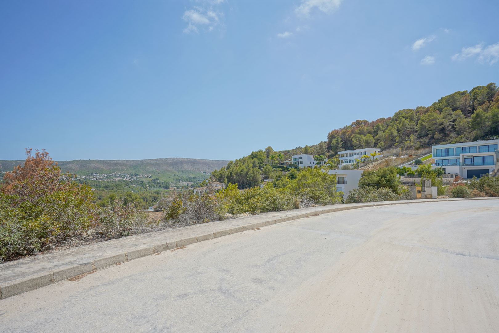 Vue depuis la route goudronnée de l'urbanisation, illustrant le terrain en pente et les villas contemporaines intégrées à la colline face à la vallée.