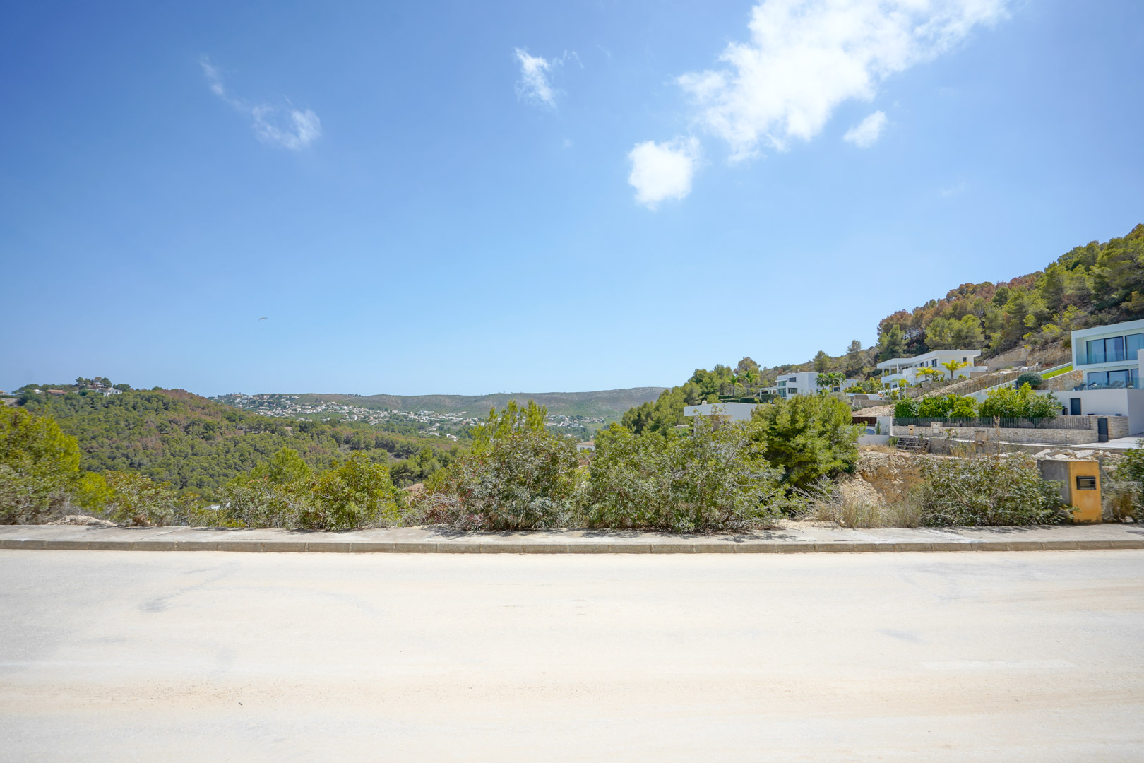 Vue panoramique depuis la route goudronnée sur les collines boisées et les villas modernes, illustrant l'infrastructure complète et l'environnement naturel.