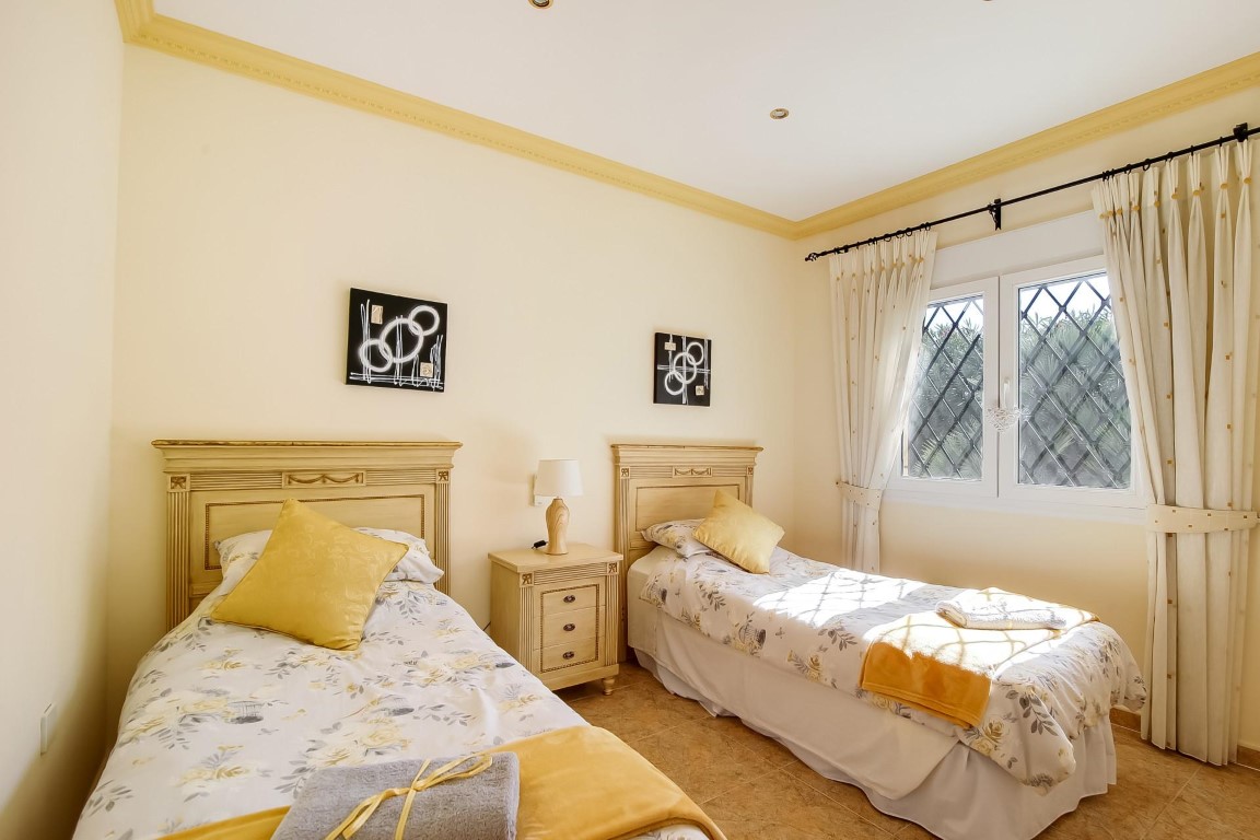 Chambre à coucher avec deux lits simples de style classique, têtes de lit en bois, moulures décoratives et fenêtre équipée de grilles de sécurité en losanges.