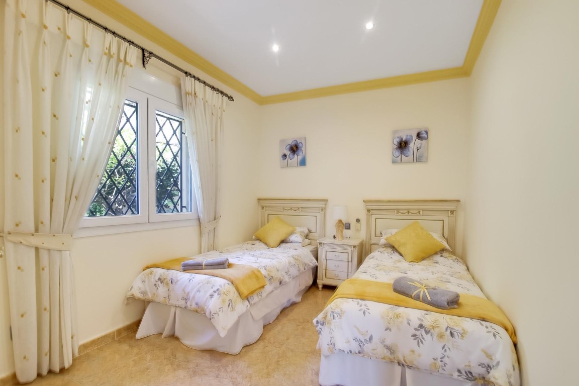Chambre à deux lits simples avec têtes de lit blanches, éclairage encastré au plafond et fenêtre sécurisée par des grilles décoratives.