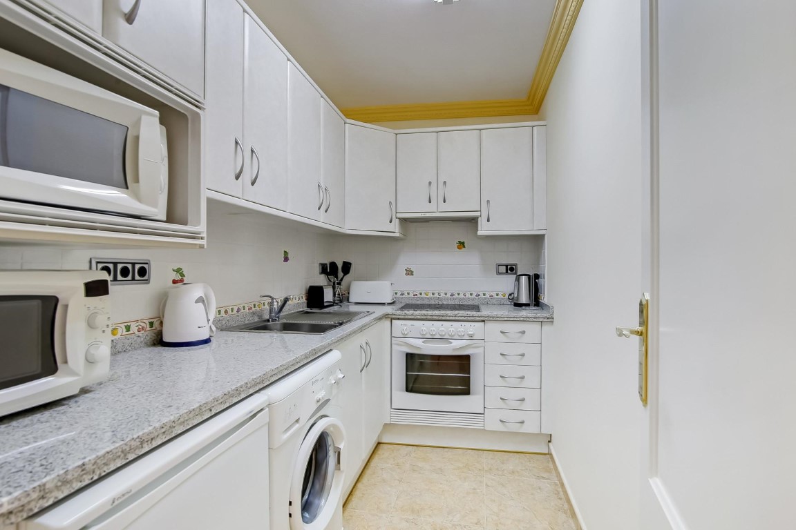 Cuisine fonctionnelle avec placards blancs, plan de travail en granit gris et électroménagers intégrés. Comprend un lave-linge et des moulures décoratives au plafond.