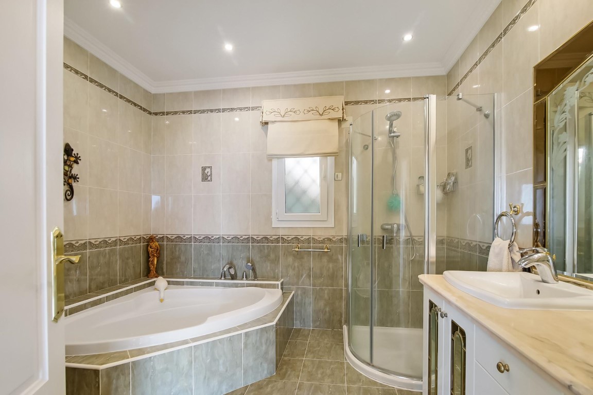Salle de bain complète avec baignoire d'angle, cabine de douche courbée et meuble vasque en marbre. Murs carrelés avec frises décoratives et éclairage encastré.