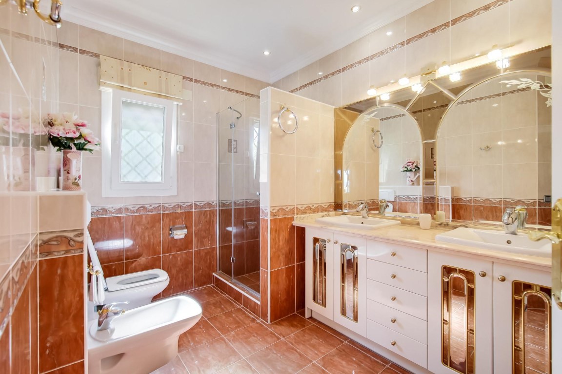 Salle de bain complète avec double vasque, miroirs cintrés à éclairage intégré, douche vitrée et carrelage aux tons terre cuite.