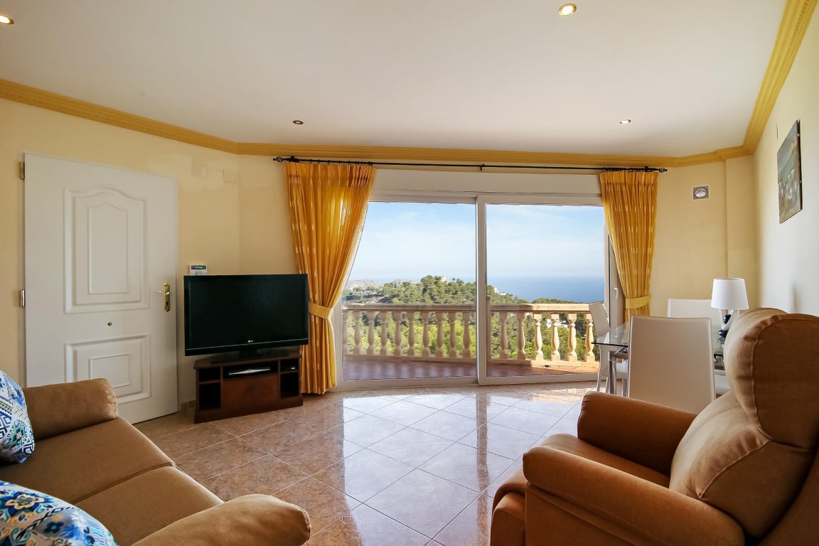 Salon lumineux avec carrelage poli, moulures au plafond et grandes baies vitrées offrant une vue panoramique sur la mer Méditerranée et les collines.