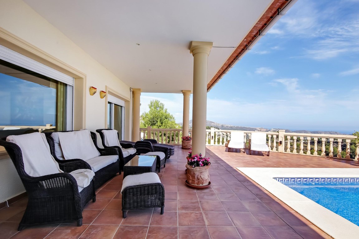 Terrasse couverte avec colonnes classiques, mobilier en osier et sol en terre cuite bordant la piscine, offrant une vue panoramique sur la mer Méditerranée.