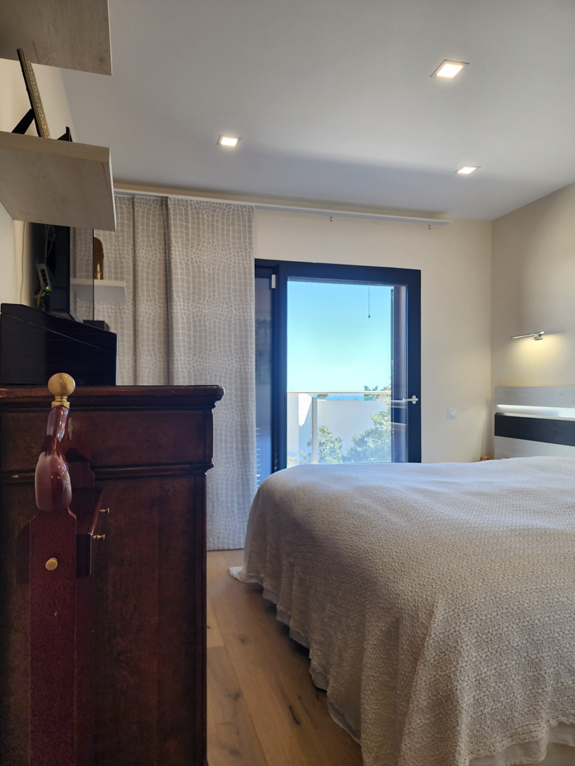 Chambre avec parquet, éclairage encastré et accès direct à la terrasse offrant une vue sur la mer via une porte vitrée à cadre sombre.