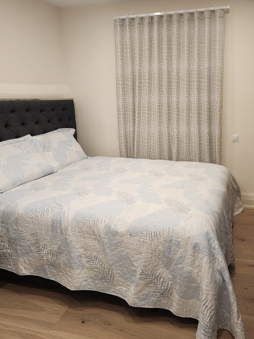 Chambre moderne avec lit double, tête de lit capitonnée gris foncé, parquet en bois clair et rideaux à motifs du plafond au sol.