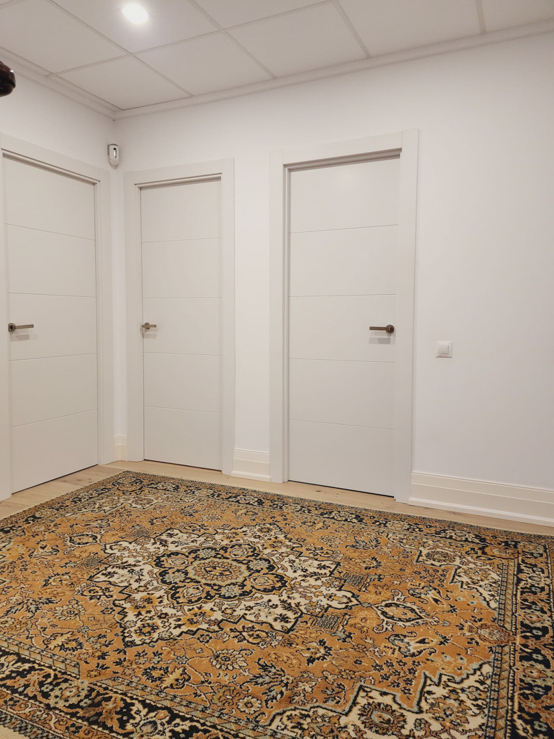Couloir de distribution avec trois portes blanches modernes, éclairage encastré et un tapis aux motifs orientaux contrastant avec les murs blancs épurés.