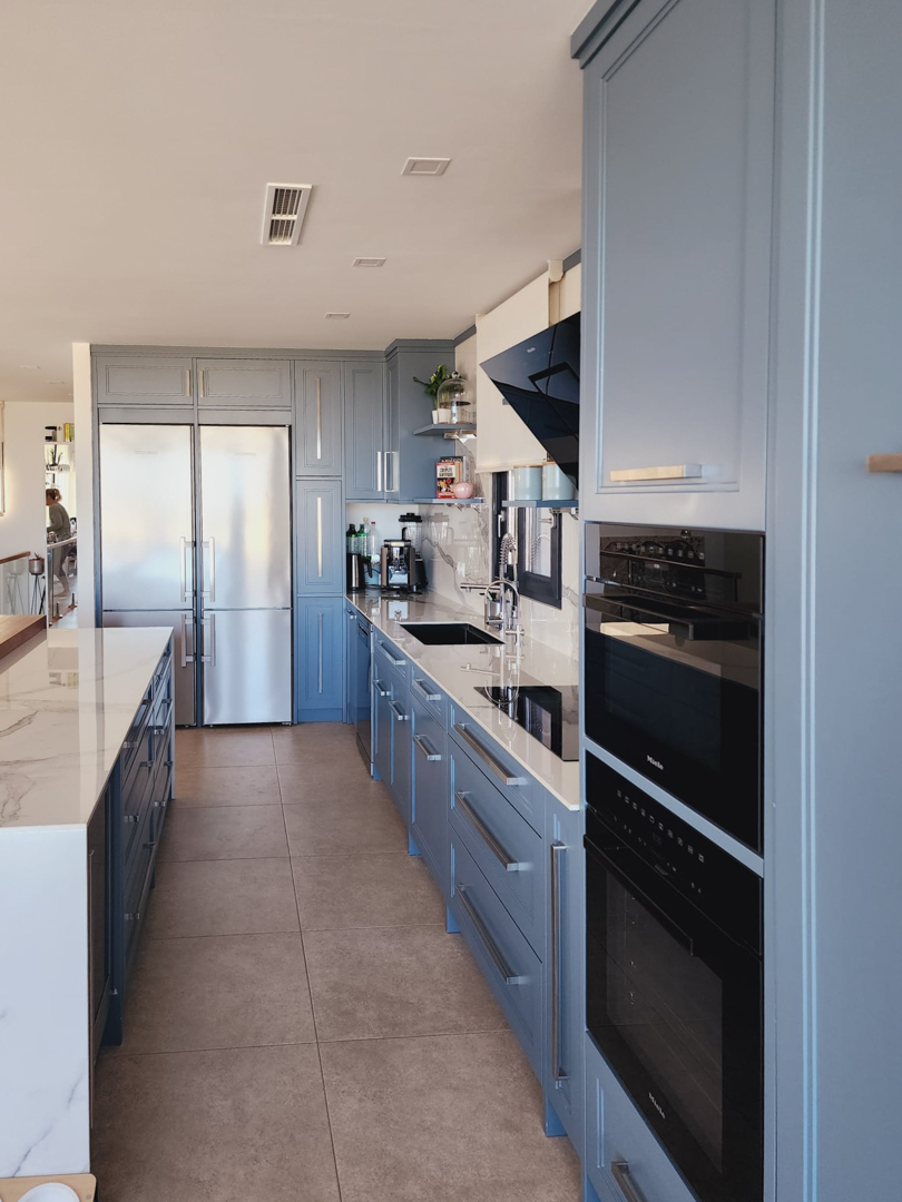 Cuisine de design avec placards bleu-gris, plans de travail effet marbre et électroménagers Miele intégrés, comprenant des fours doubles et un grand réfrigérateur en inox.