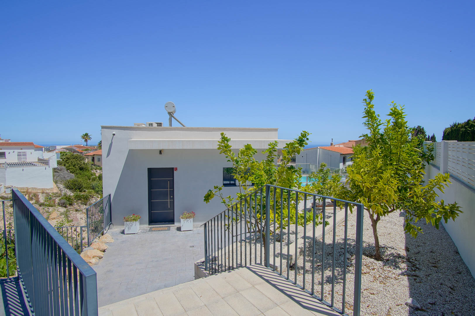 Entrée principale de la villa avec porte grise, façade blanche épurée, chauffe-eau solaire et jardin méditerranéen composé de graviers et d'agrumes.