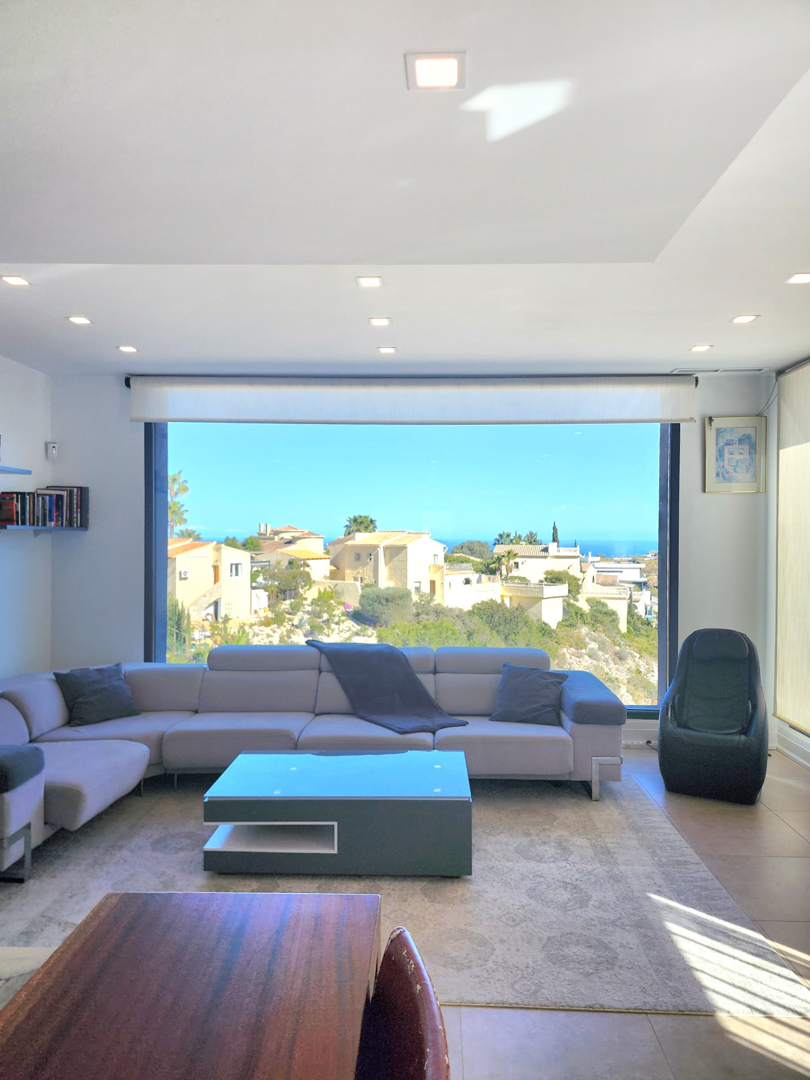 Espace de vie lumineux avec une grande fenêtre panoramique sur la mer, un canapé d'angle gris et un éclairage de plafond encastré moderne.