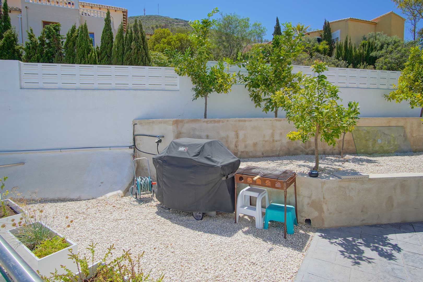Espace extérieur avec gravier blanc, arbres fruitiers et coin barbecue. Le jardin est délimité par un mur blanc offrant une vue sur les collines voisines.