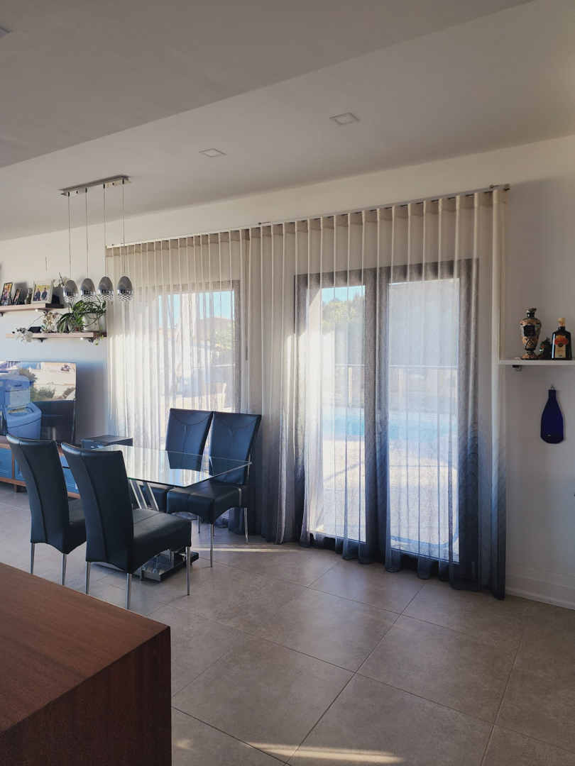 Espace repas lumineux avec table en verre et chaises à dossier haut, situé devant de grandes baies vitrées avec rideaux dégradés menant à la terrasse de la piscine.