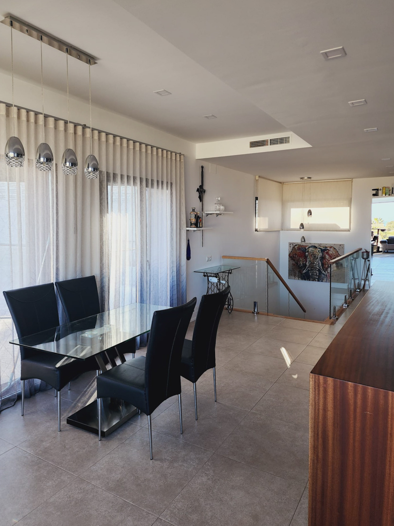 Espace salle à manger avec table en verre et chaises noires, bordé par un escalier avec garde-corps vitré. L'éclairage est assuré par des suspensions chromées et des spots intégrés.