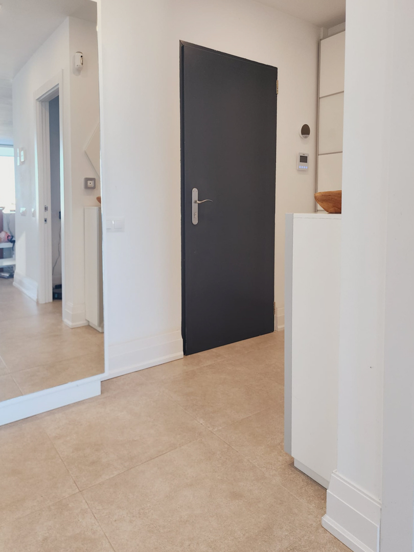 Hall d'entrée avec carrelage clair, grand miroir mural et porte sombre minimaliste, intégrant des commandes domotiques et des finitions soignées.