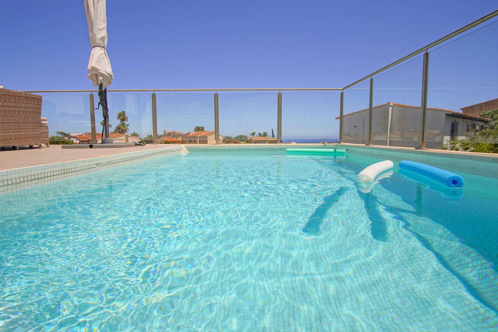 Piscine rectangulaire avec mosaïque bleu clair et garde-corps en verre. La terrasse ensoleillée dispose de mobilier de jardin et offre une vue dégagée sur l'horizon marin.