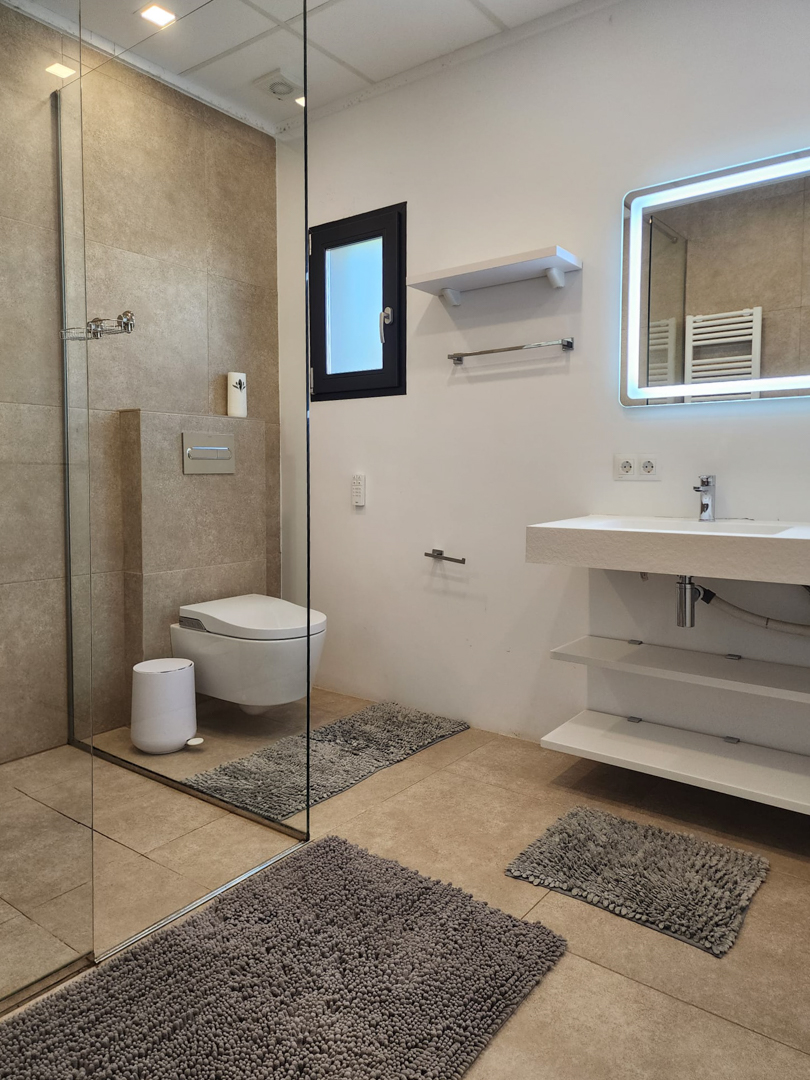 Salle de bain moderne avec douche à l'italienne, paroi vitrée, WC suspendu et meuble vasque à étagères ouvertes complété par un miroir rétroéclairé.