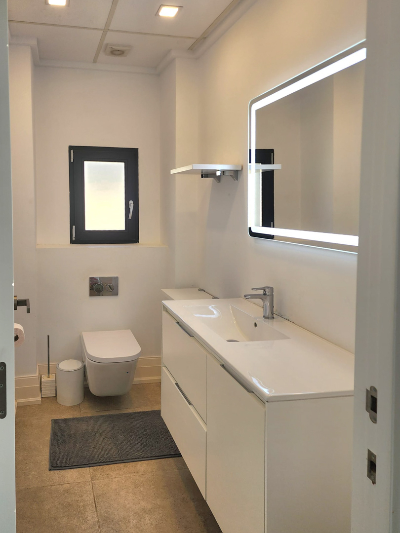 Salle de bain moderne avec meuble vasque blanc, miroir rétroéclairé par LED et WC suspendu. Finitions épurées avec éclairage encastré et carrelage au sol neutre.
