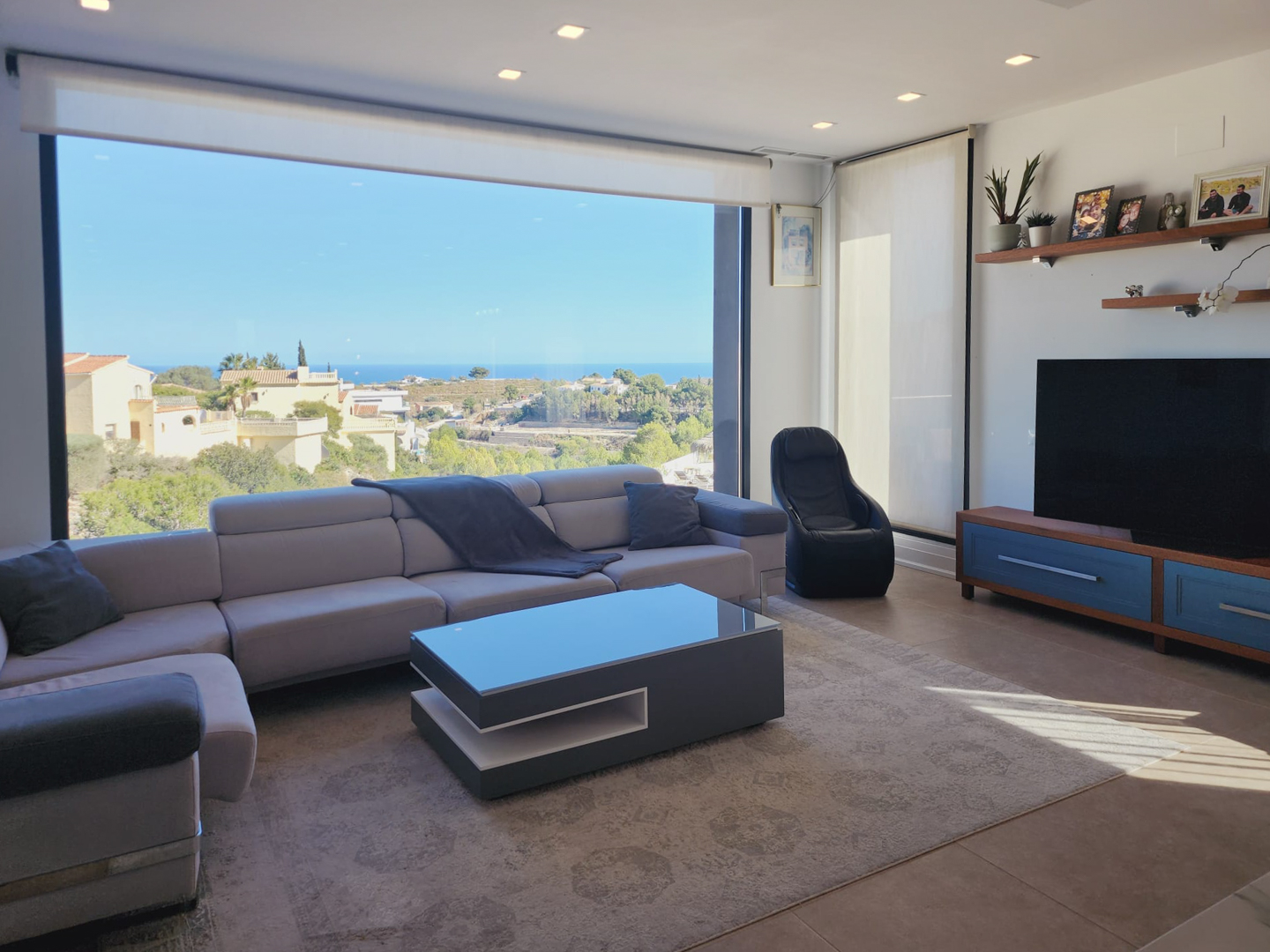 Salon moderne avec grande baie vitrée offrant une vue sur la mer, canapé d'angle gris, table basse design et éclairage par spots intégrés.