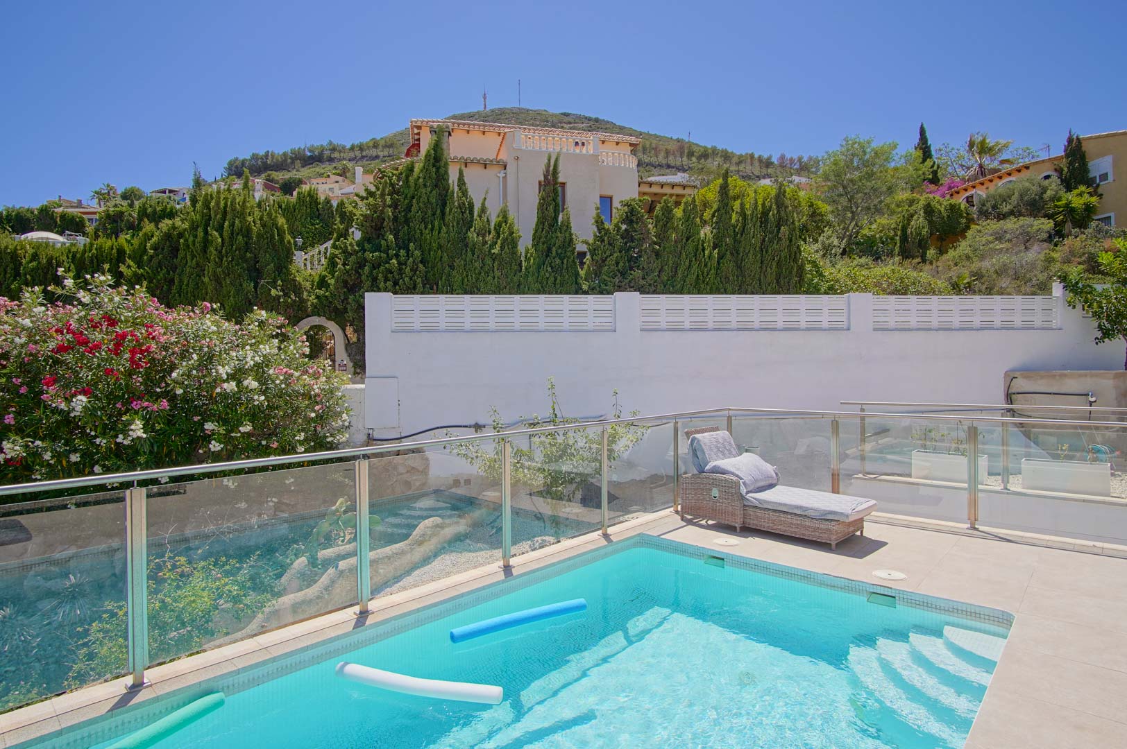 Terrasse ensoleillée avec piscine rectangulaire, balustrades en verre et transat en osier, bordée de lauriers-roses et de cyprès verdoyants.