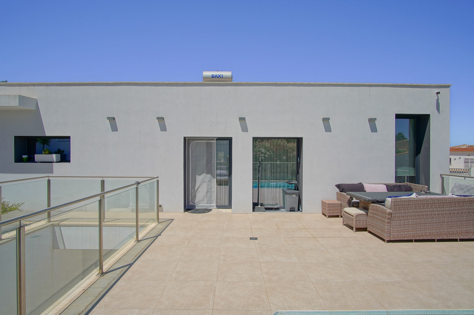 Terrasse supérieure avec carrelage en grès cérame, mobilier d'extérieur en rotin et garde-corps en verre. Chauffe-eau solaire BAXI installé sur le toit plat de la villa.