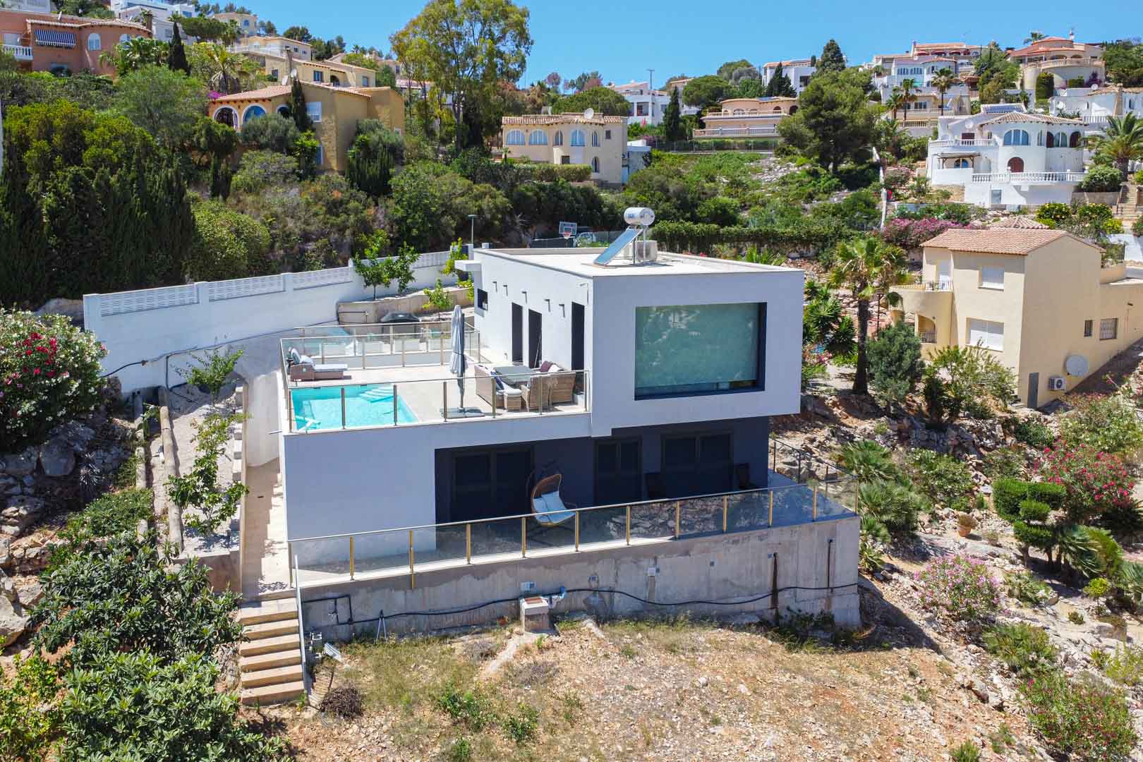 Vue extérieure de la villa moderne au design cubique, avec une terrasse supérieure comprenant piscine, garde-corps en verre et chauffe-eau solaire sur le toit.