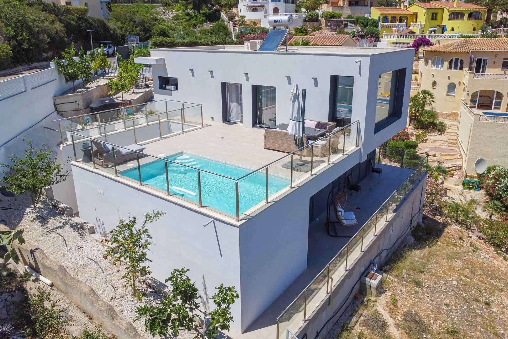 Vue extérieure de la villa moderne avec piscine sur la terrasse supérieure, garde-corps en verre, panneaux solaires et terrasse couverte au niveau inférieur.