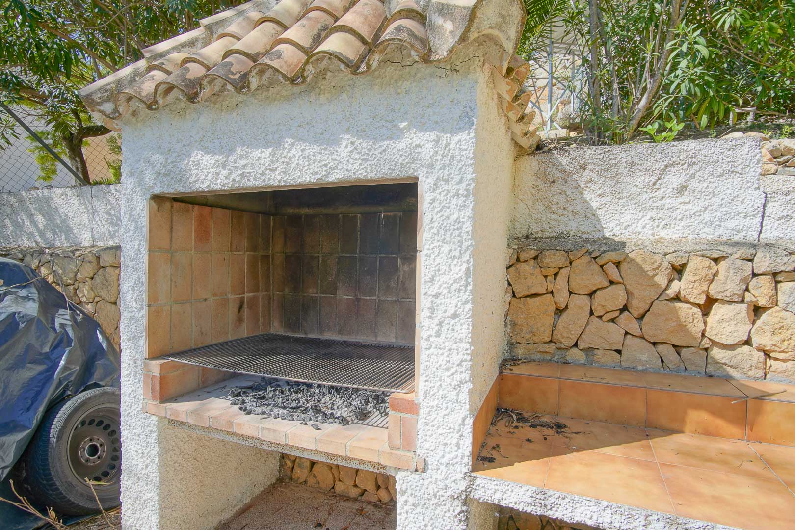 Barbecue maçonné avec toit en tuiles de terre cuite et finition en crépi blanc, installé contre un mur en pierre naturelle avec un plan de travail carrelé.