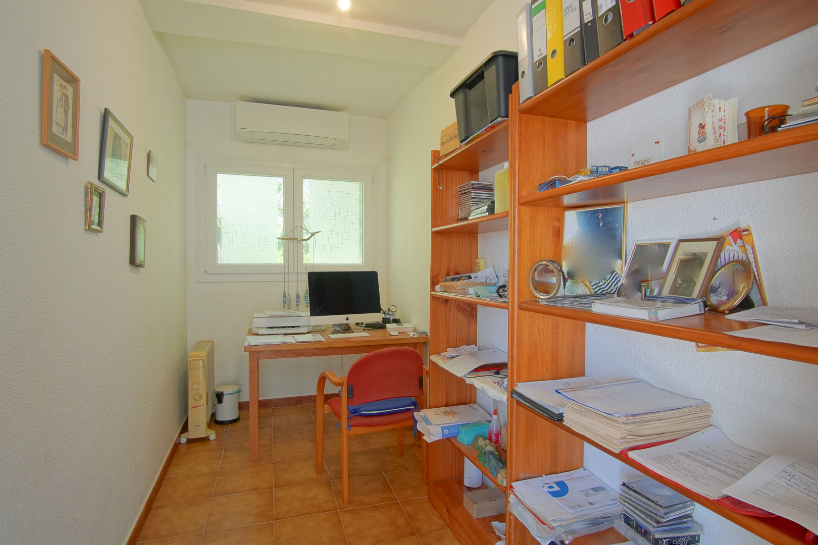 Bureau fonctionnel avec bureau en bois, étagères de rangement, sol en grès cérame et climatisation murale, offrant un espace de travail calme.