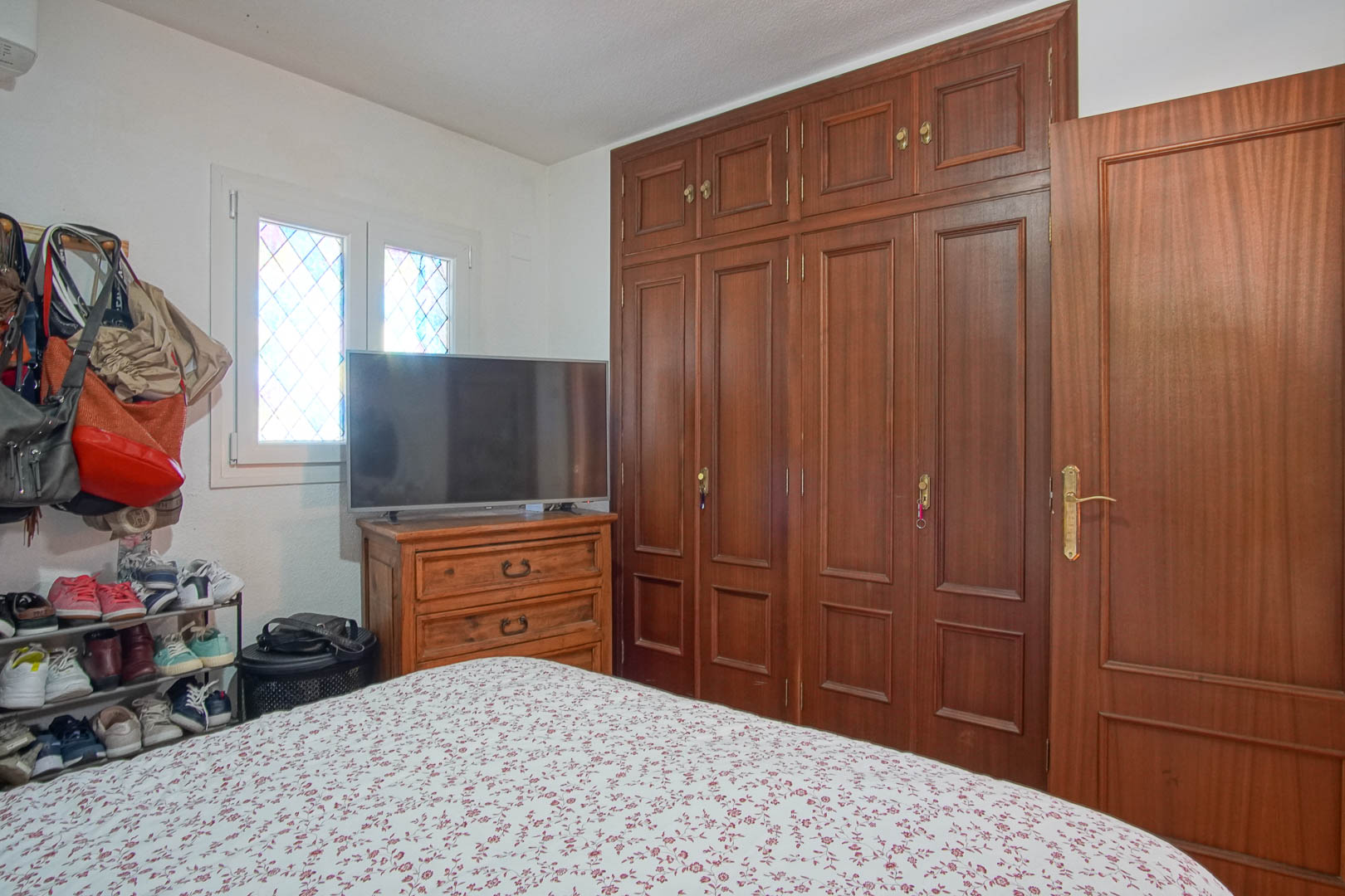 Chambre avec grand placard intégré en bois sombre, commode assortie surmontée d'un téléviseur et fenêtre aux vitres plombées artisanales.