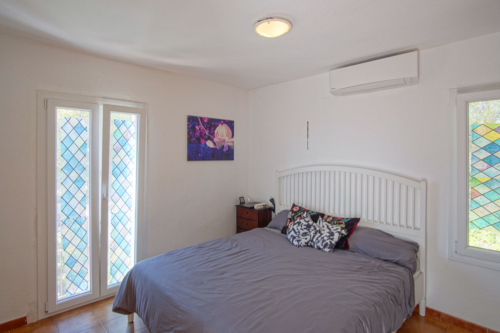 Chambre lumineuse avec climatisation réversible, lit double à tête de lit blanche et fenêtres ornées de vitraux géométriques colorés.