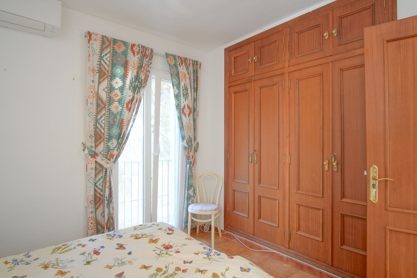 Chambre lumineuse avec de grands placards intégrés en bois, climatisation murale et accès extérieur par une porte-fenêtre ornée de rideaux à motifs géométriques.