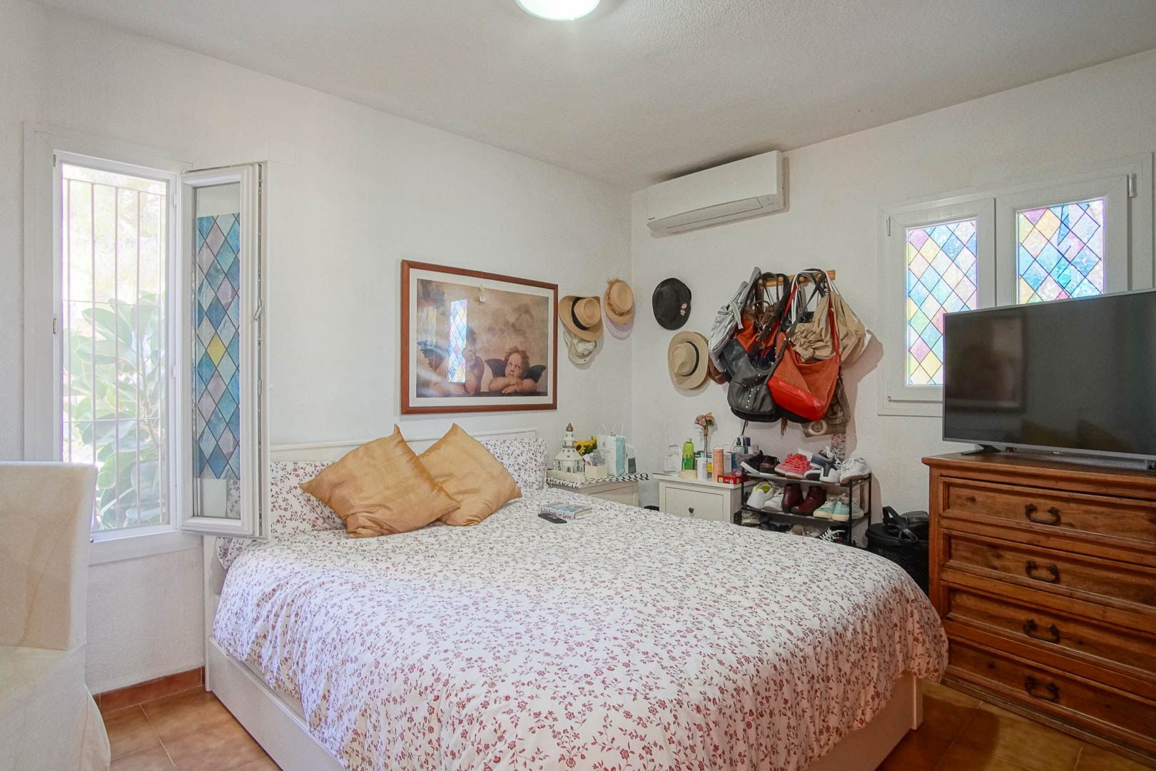 Chambre lumineuse avec sol en grès, climatisation et fenêtres ornées de vitraux colorés. Elle dispose d'une commode en bois classique et d'un bel éclairage naturel.