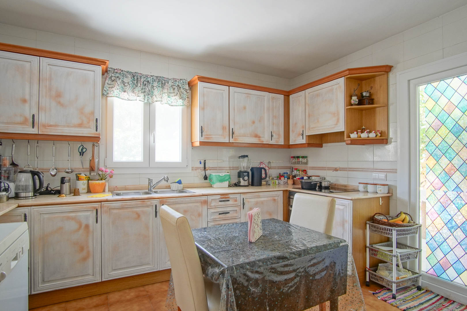 Cuisine indépendante de style rustique avec des meubles à l'aspect vieilli, un plan de travail en L et un coin repas. Une porte vitrée aux motifs colorés apporte une luminosité unique.