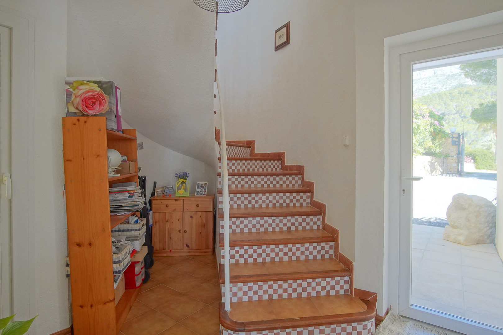 Escalier intérieur avec marches en terre cuite et contremarches à damier, mobilier en pin et porte vitrée ouvrant sur l'extérieur ensoleillé.