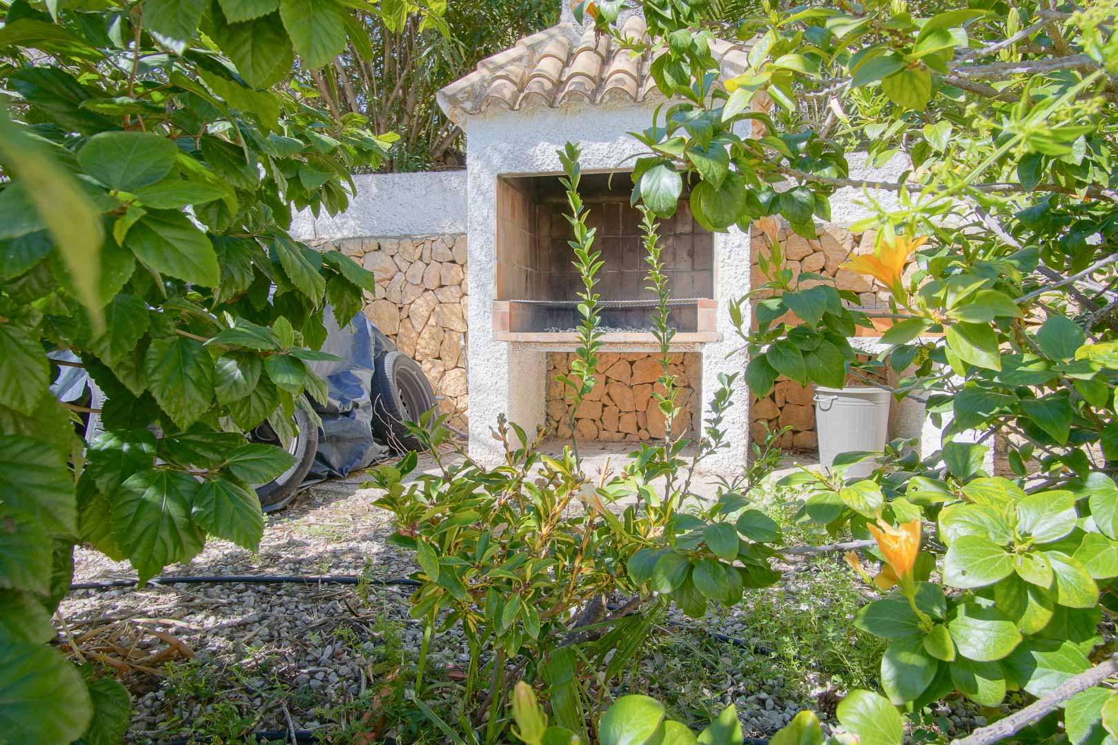 Espace barbecue maçonné avec toit en tuiles, niché dans un jardin verdoyant bordé de murs en pierre naturelle apparente.