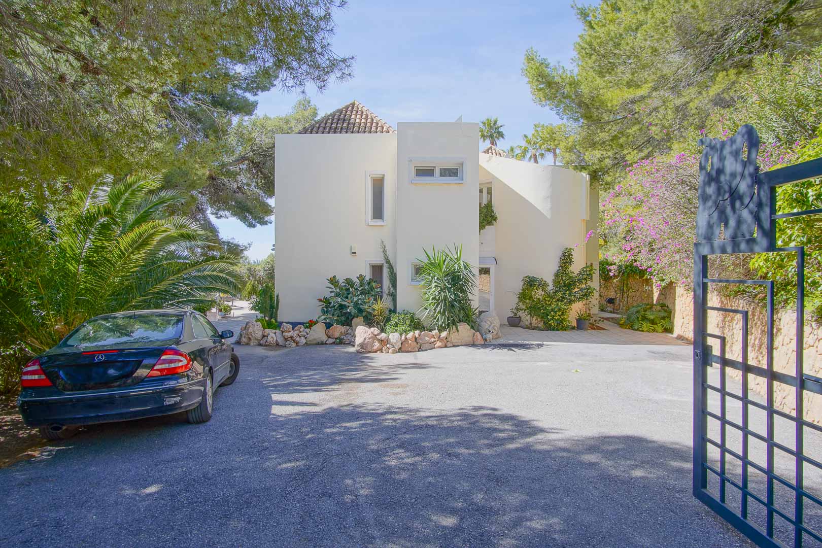 Façade principale de la villa à l'architecture méditerranéenne, avec un grand espace de stationnement pavé et une entrée sécurisée entourée de pins et de végétation.