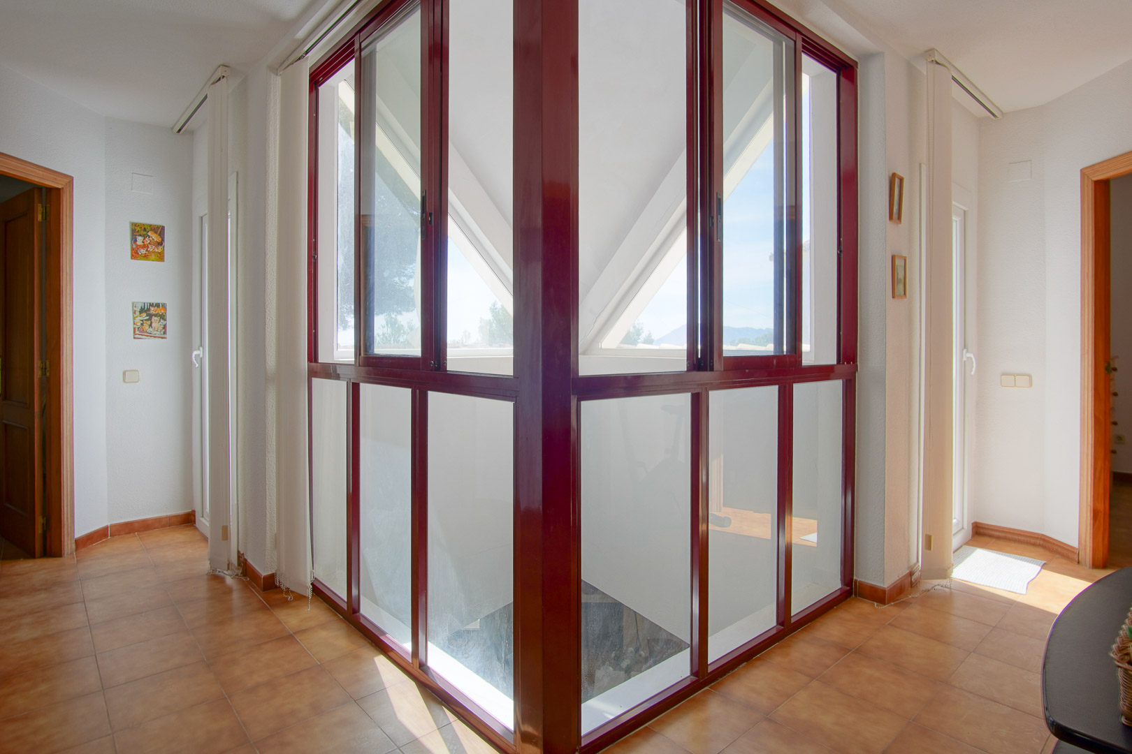 Palier à l'étage avec une structure vitrée d'angle aux cadres bordeaux, carrelage en terre cuite et une luminosité naturelle reliant les différentes pièces.