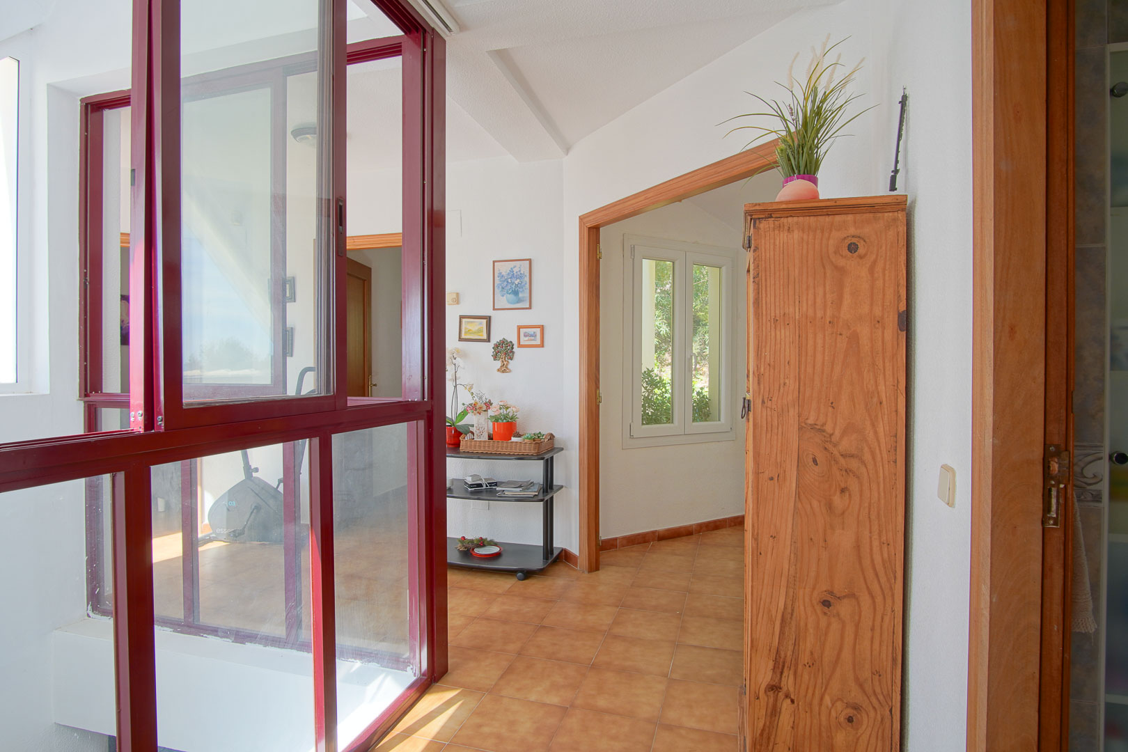 Palier lumineux avec sol en grès cérame, encadrements en bois et une cloison vitrée aux châssis bordeaux séparant un espace de fitness intérieur.