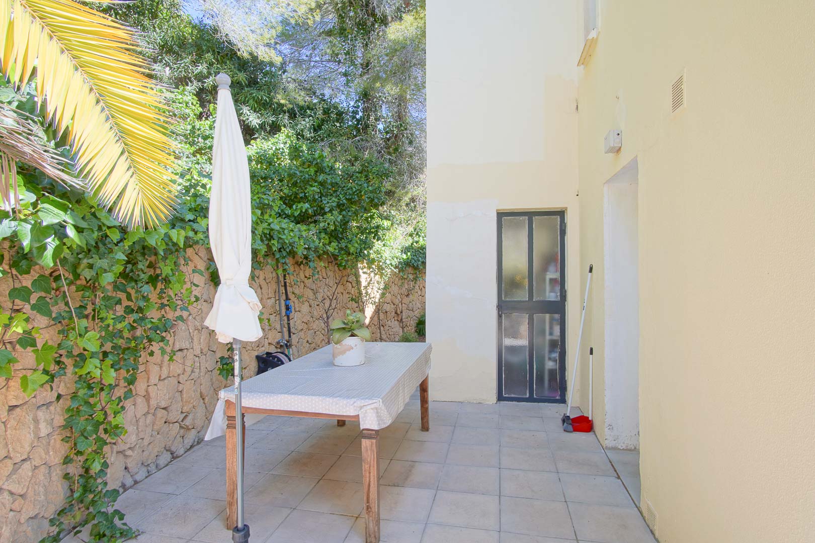 Patio extérieur pavé bordé d'un mur en pierre naturelle et de lierre, comprenant une table à manger et un parasol dans un cadre verdoyant et calme.