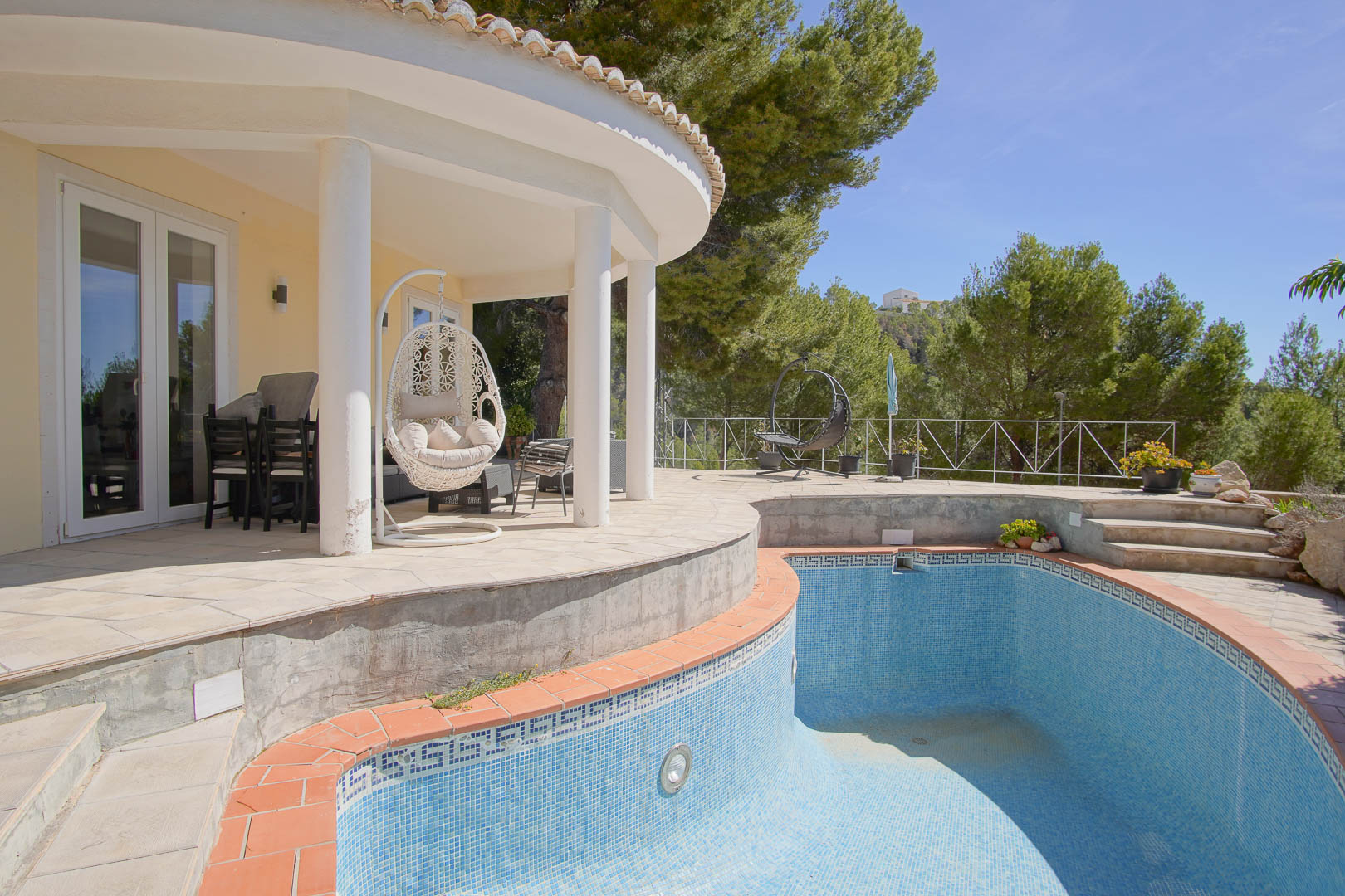 Porche couvert de forme arrondie avec colonnes blanches et fauteuil suspendu, offrant une vue sur la piscine en mosaïque et la pinède environnante.