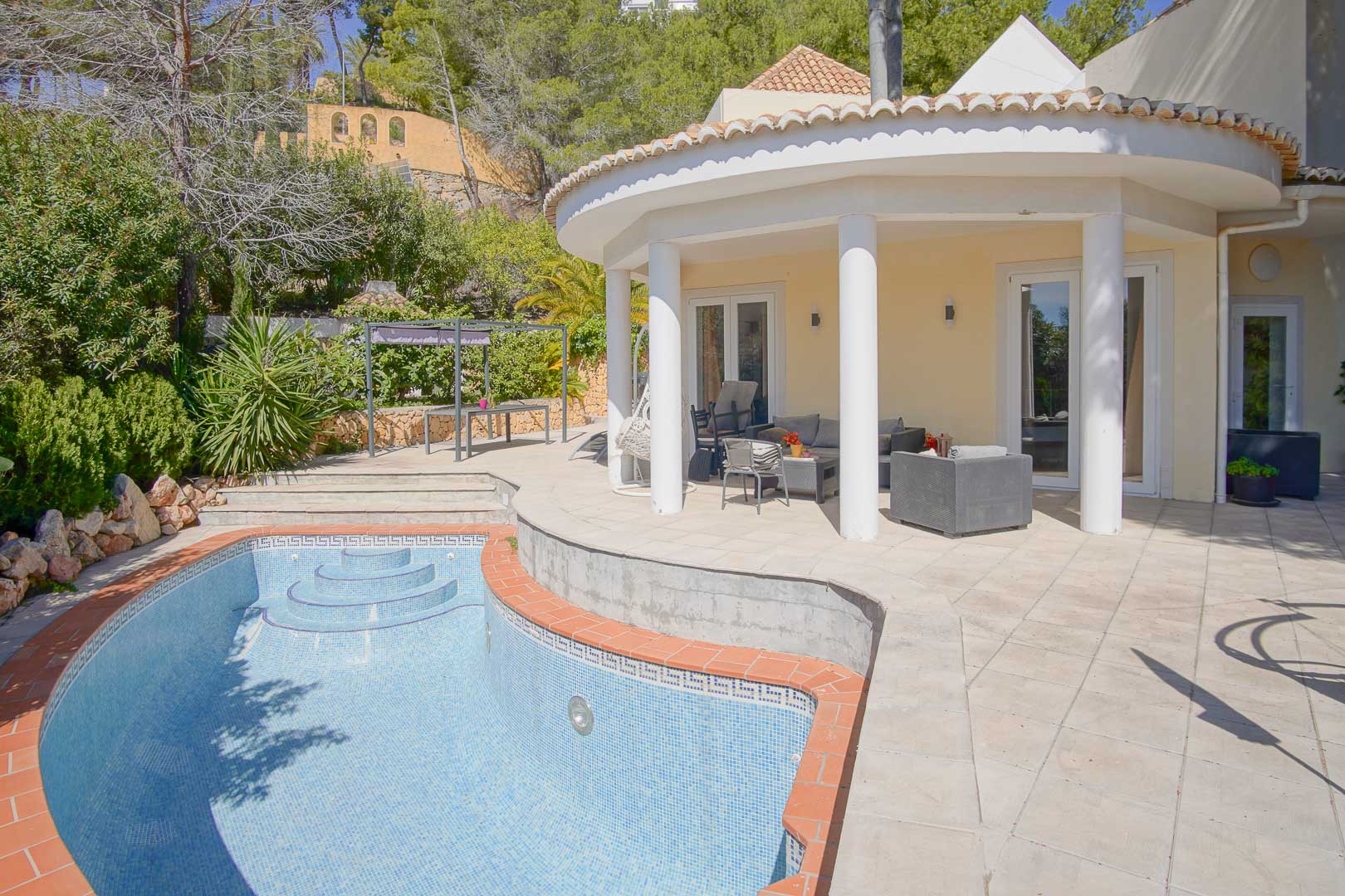 Porche semi-circulaire avec colonnes blanches donnant sur une piscine privée et une terrasse en pierre, entouré d'un jardin méditerranéen arboré.