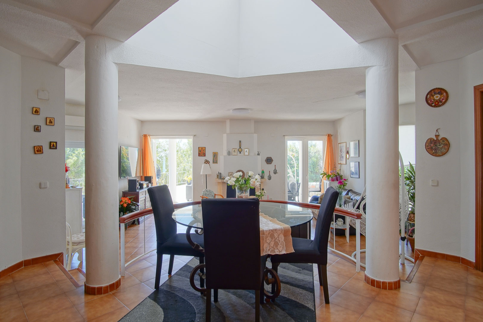 Salle à manger lumineuse sous un puits de lumière central, avec colonnes blanches, sol en terre cuite et vue sur le salon avec cheminée.