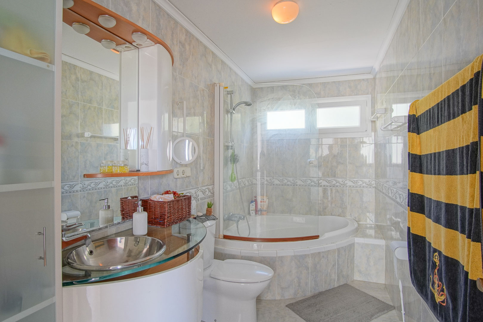 Salle de bain complète avec baignoire d'angle, paroi en verre et vasque moderne en inox sur plan de travail vitré. Murs carrelés avec frise décorative et lumière naturelle.