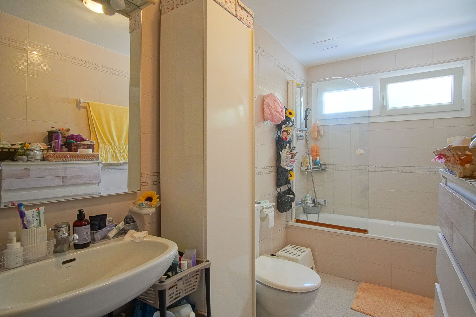 Salle de bain complète avec baignoire, paroi vitrée, lavabo blanc et armoire colonne. Carrelage mural beige et fenêtre assurant une luminosité naturelle.