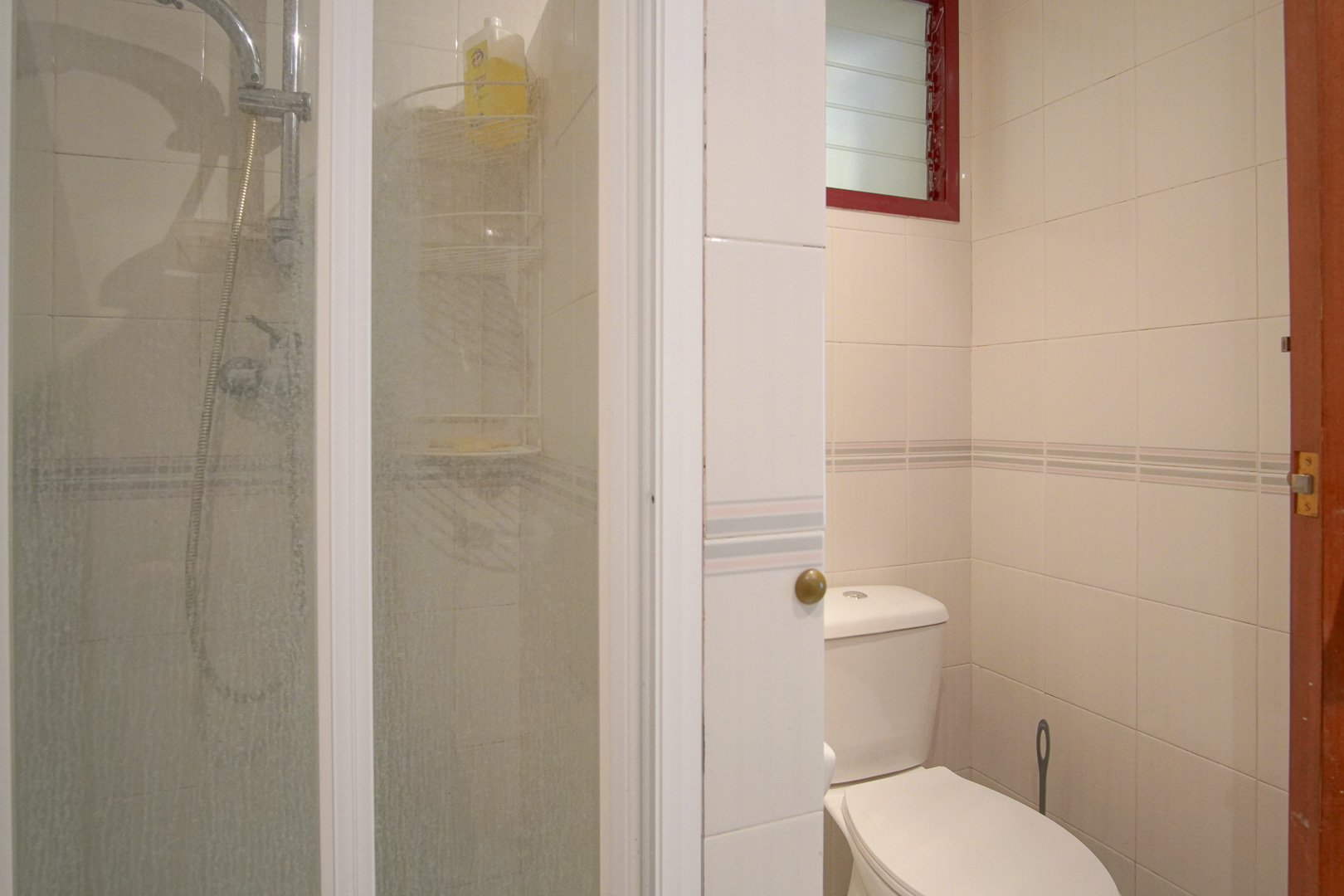 Salle de bain fonctionnelle avec murs carrelés en blanc, douche avec paroi vitrée, toilettes et petite fenêtre de ventilation à cadre rouge.