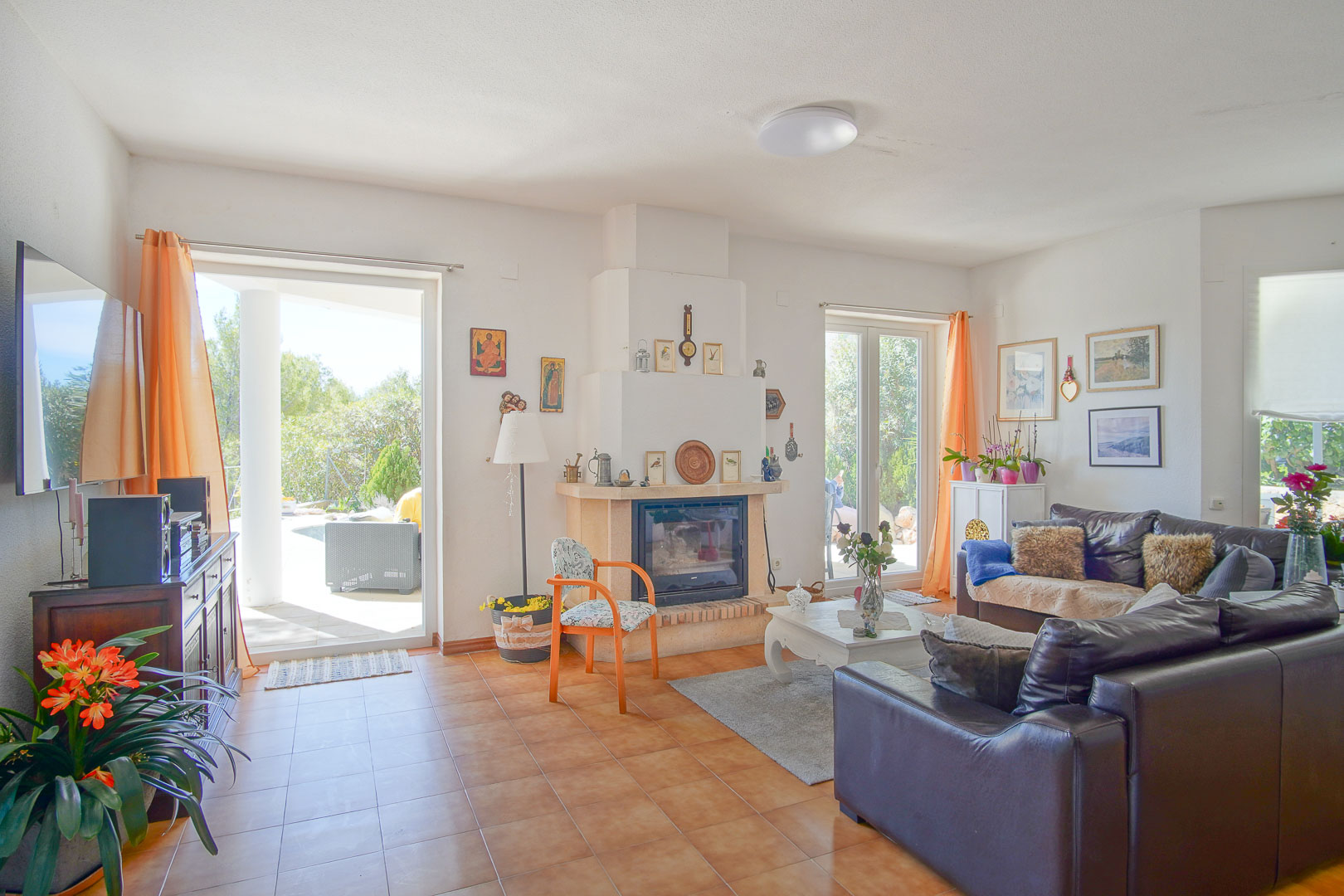 Salon lumineux avec cheminée centrale, sol en terre cuite et accès direct au porche extérieur par de grandes baies vitrées.
