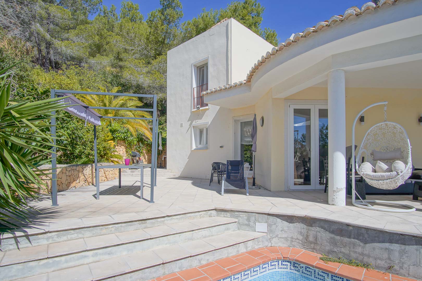 Terrasse extérieure avec bord de piscine, espace repas sous pergola et porche couvert avec fauteuil suspendu, entourée d'une végétation méditerranéenne.